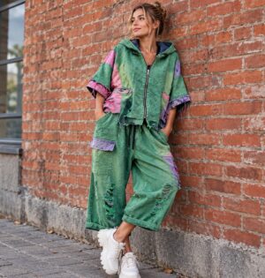 Komplet damski patchwork, kurtka z kapturem i szerokie spodnie - sesja miejska: modelka w zielonym komplecie, ceglany mur, sneakersy na grubej podeszwie, usunięte logo