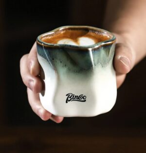 Kubek ceramiczny Bincoo 180 ml bez ucha, nieregularny kształt, do kawy - dłoń trzymająca kubek z cappuccino przy oknie kawiarni