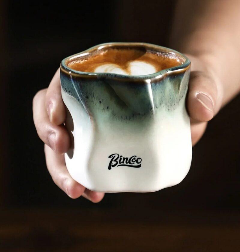 Kubek ceramiczny Bincoo 180 ml bez ucha, nieregularny kształt, do kawy - dłoń trzymająca kubek z cappuccino przy oknie kawiarni