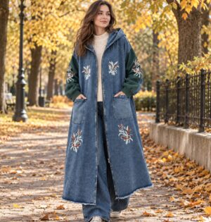 Płaszcz jeansowy damski z kapturem i haftem w kwiaty, długi, oversize - jesienny park, modelka w niebieskim płaszczu, kremowy sweter i białe trampki